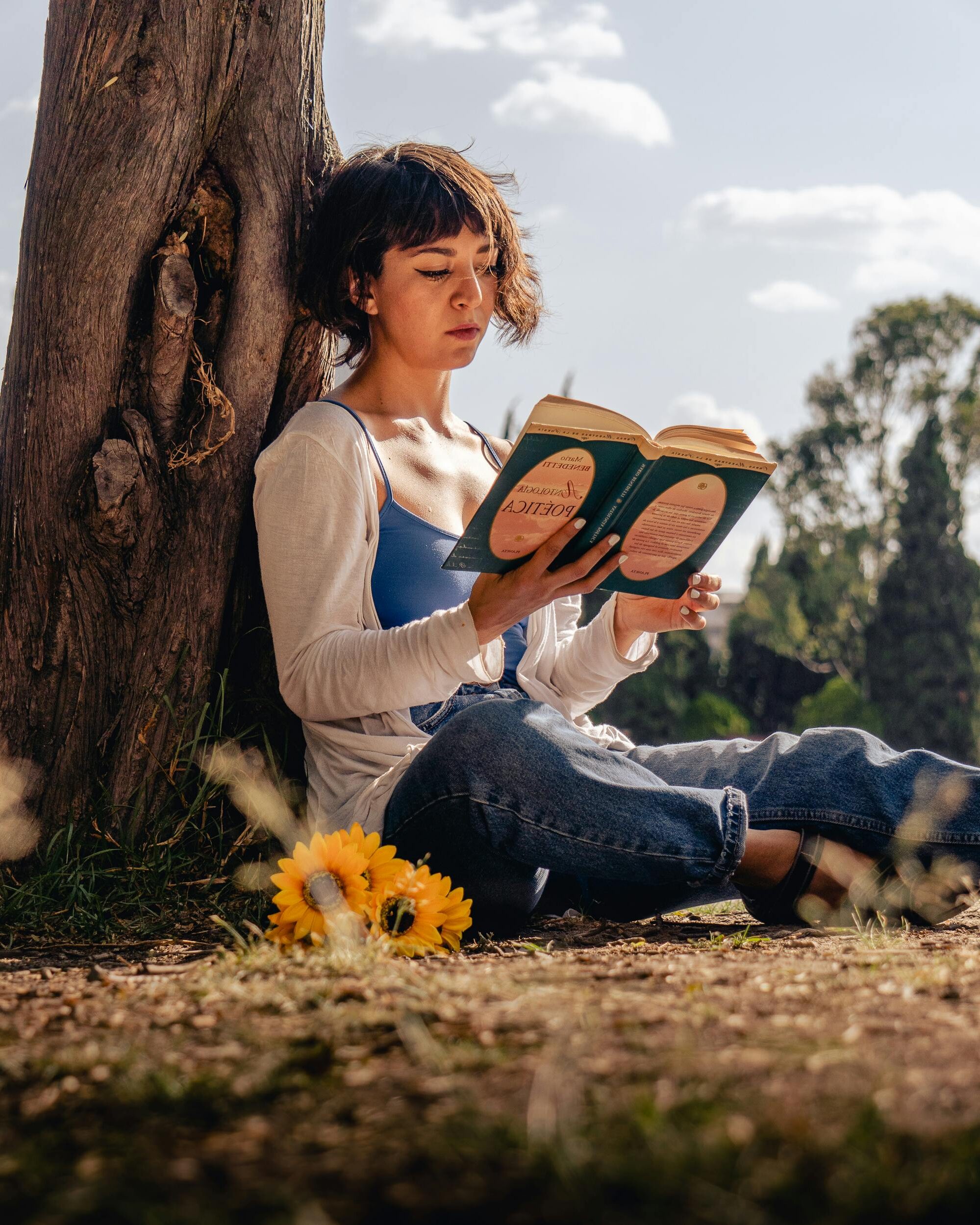 Young woman reading a book while sitting against a tree with flowers in Mexico City park.